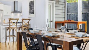 Un comedor con una mesa de madera dispuesta para cuatro personas, rodeado de sillas negras. Se ve una cocina al fondo con armarios blancos y una campana extractora de acero inoxidable.