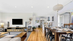 Una sala de estar moderna con televisor de pantalla plana, piso de madera, una zona de comedor con mesa y sillas de madera, y una cocina al fondo.