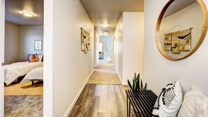 Un couloir avec un plancher en bois, un lit avec une literie blanche, un miroir avec un cadre en bois et une plante.