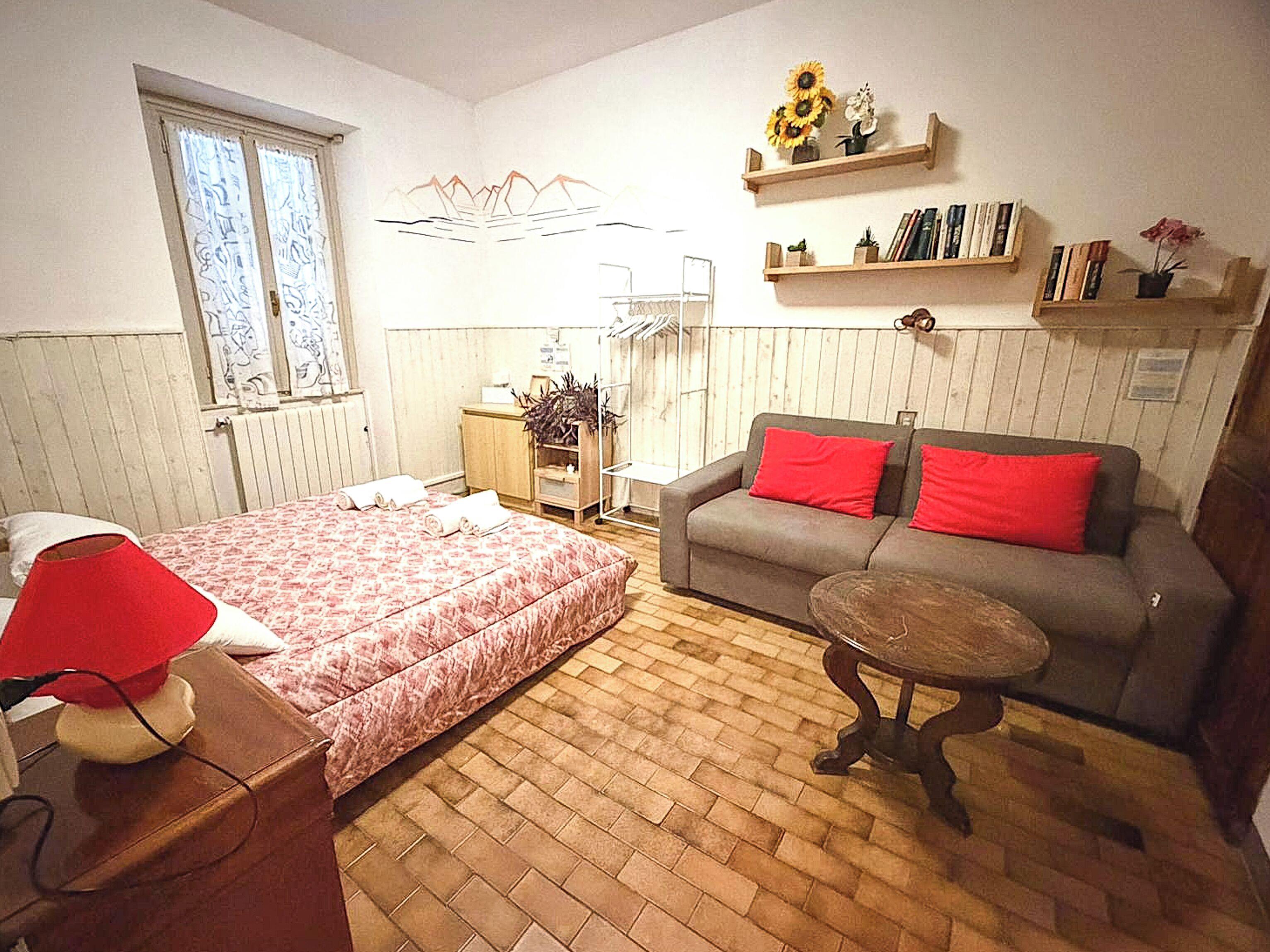 Un dormitorio con cama, sofá, una mesita, radiador, ventana y estanterías con libros y flores.
