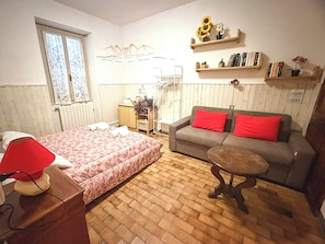 Quarto com cama, sofá, mesa pequena, radiador, janela e prateleiras com livros e flores.