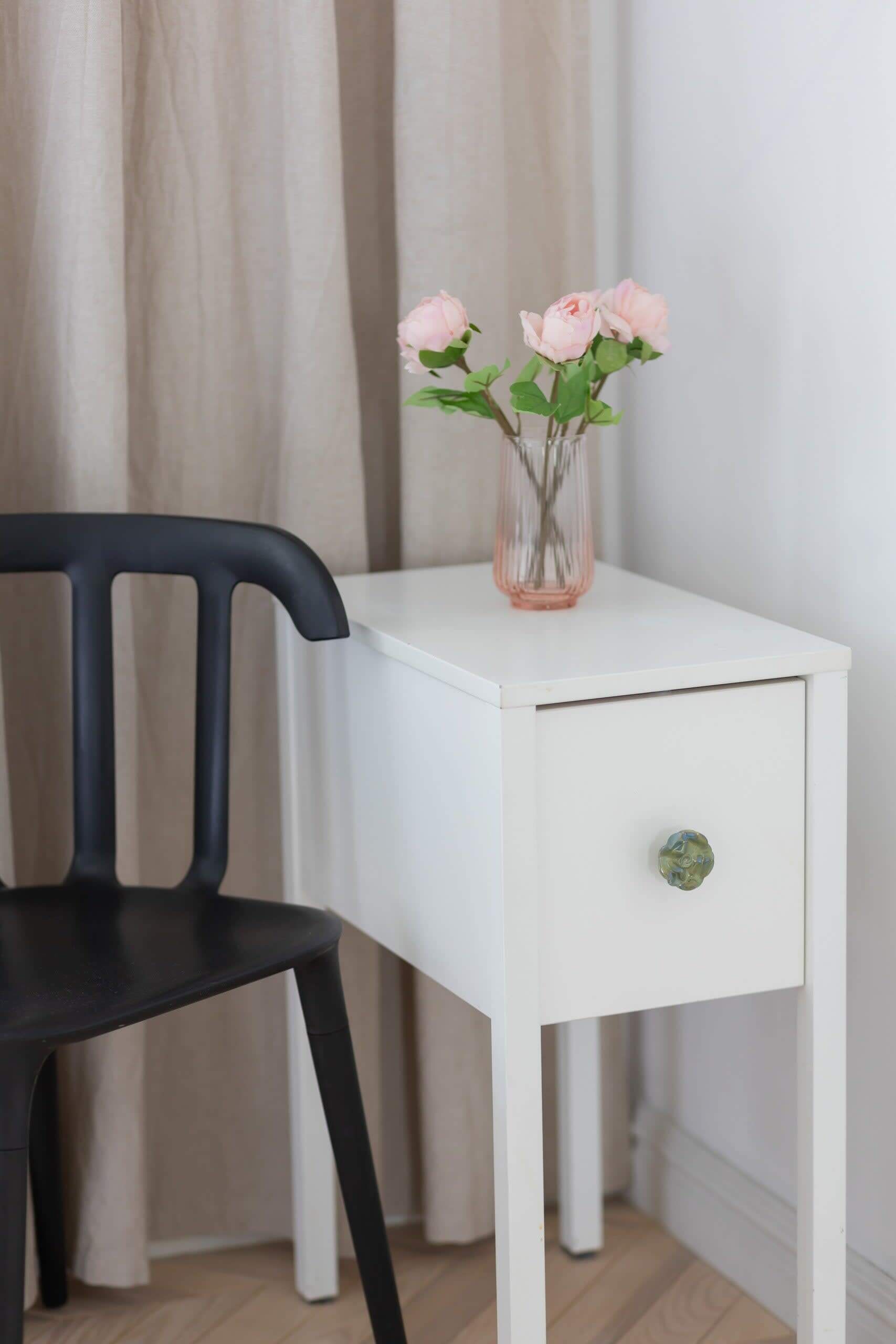 A white bedside table with a drawer, a vase with pink flowers, and a black chair.