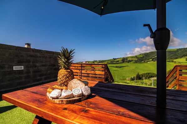 A wooden table with a pineapple, cheese, and bread on a sunny patio with a view of green hills.