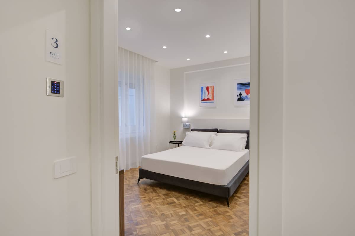 A modern hotel room with a large bed, a bedside table, a wall-mounted light, and a window with sheer curtains.