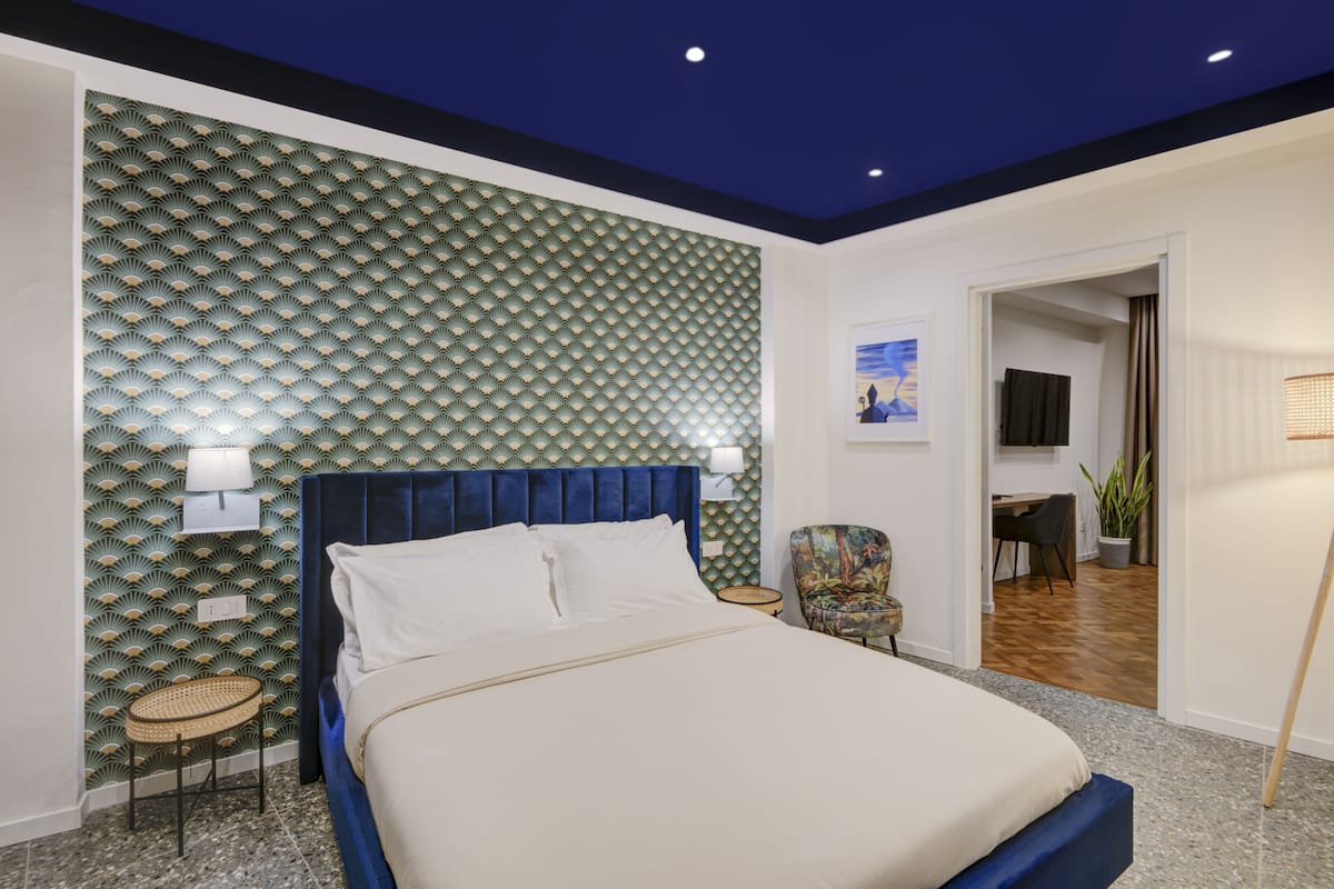 A modern bedroom with a blue headboard, a bed with white linens, a wall with a green patterned design, a small round table with a lamp, and a chair with a colorful pattern.
