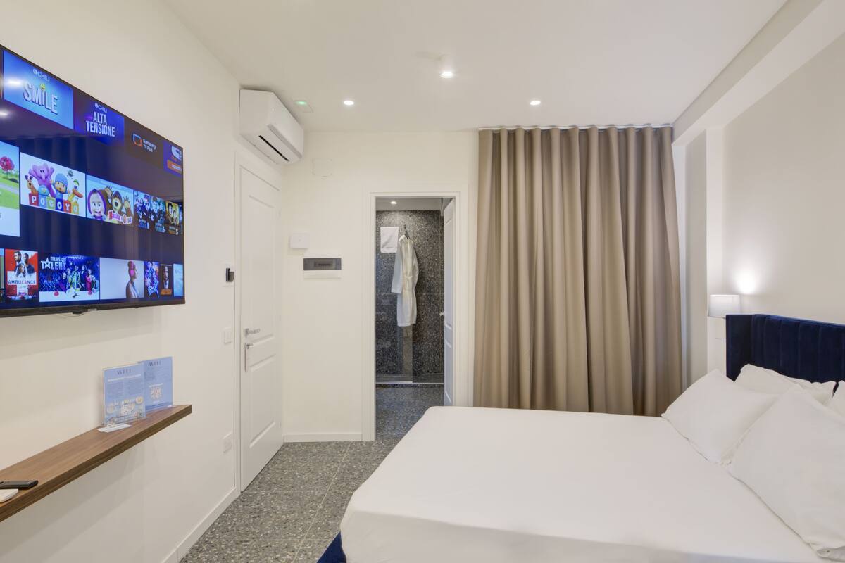 A hotel room with a bed, a TV mounted on the wall displaying various channels, a shower area, and a curtain.