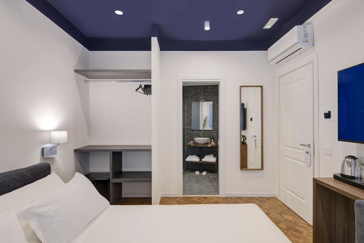 A modern hotel room with a bed, a desk, a mirror, a TV, and a bathroom visible through an open door.