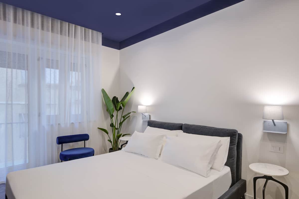 A modern hotel room with a large bed, a small table, a chair, and a potted plant.