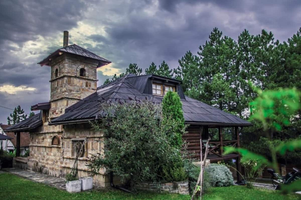 Un edificio rústico de piedra con una torre, un porche cubierto y un jardín bien cuidado.