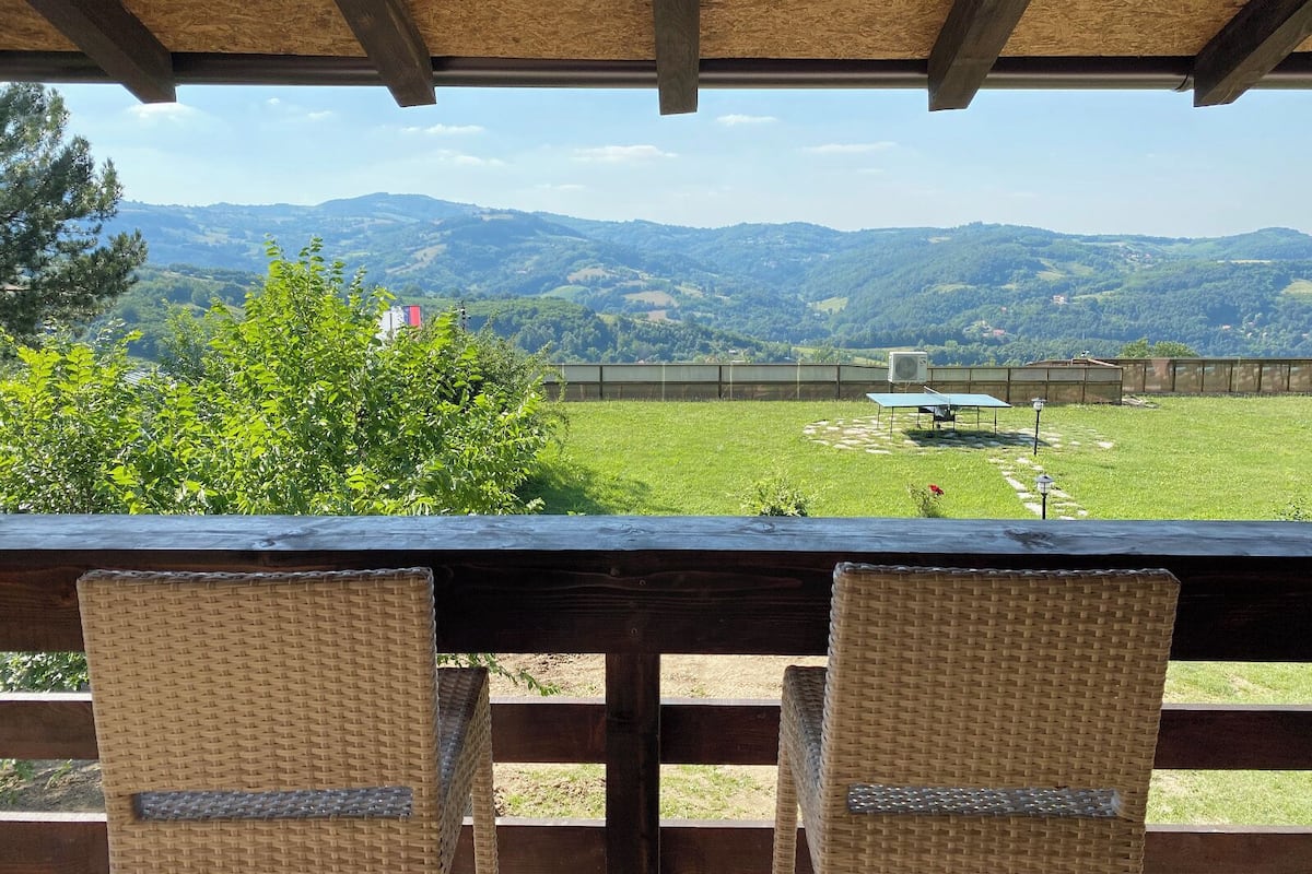 Un patio con sillas de mimbre, una mesa y vistas a un campo verde y montañas.