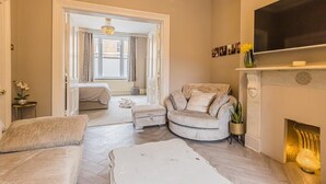 A spacious living room with a fireplace, a television mounted above it, and a comfortable seating area with a sofa and ottoman. - Ground Floor 1 Bedroom Flat - Windsor By Tempstay (Berkshire)