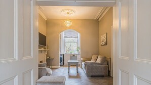 A hallway leading to a living room with a chandelier, a sofa, a table, and a window with a view. - Ground Floor 1 Bedroom Flat - Windsor By Tempstay (Berkshire)