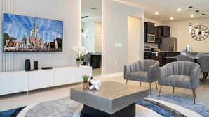 Ein modernes Wohnzimmer mit einem Flachbildfernseher, grauen Sofas, einem Couchtisch und einer Küche im Hintergrund.