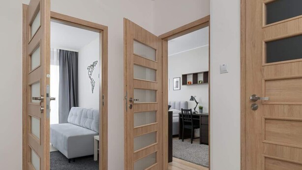 A modern hotel room with a grey sofa, wooden doors with glass panels, a dining area with a table and chairs, and a small kitchen area.