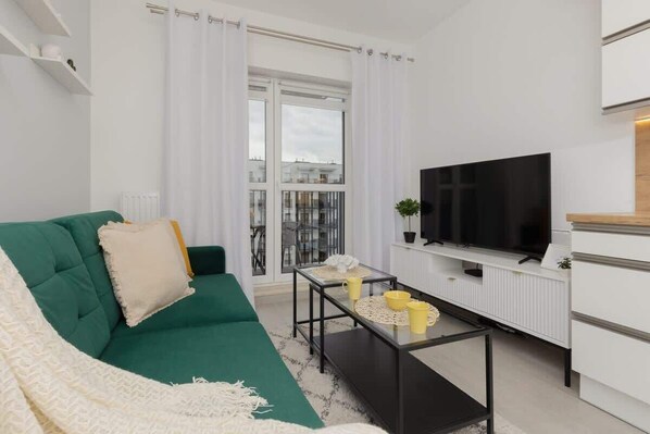 A modern living room with a green sofa, a flat-screen TV, a small coffee table, and a view of the city through large windows.