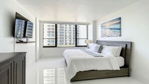 A modern bedroom with a large bed, a flat-screen TV, a window with a city view, and a painting on the wall. - Deluxe 1-bed 2-bath ocean front free valet parking on the beach (Hollywood)