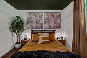 Une chambre à coucher avec un lit, deux tableaux abstraits accrochés au mur, une plante en pot et une table de chevet avec une lampe.