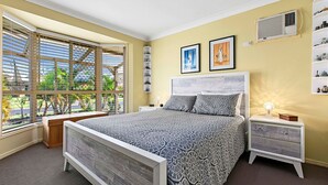 Un dormitorio con una cama grande, una ventana con persianas, una mesita de noche con lámpara y un estante en la pared con adornos.