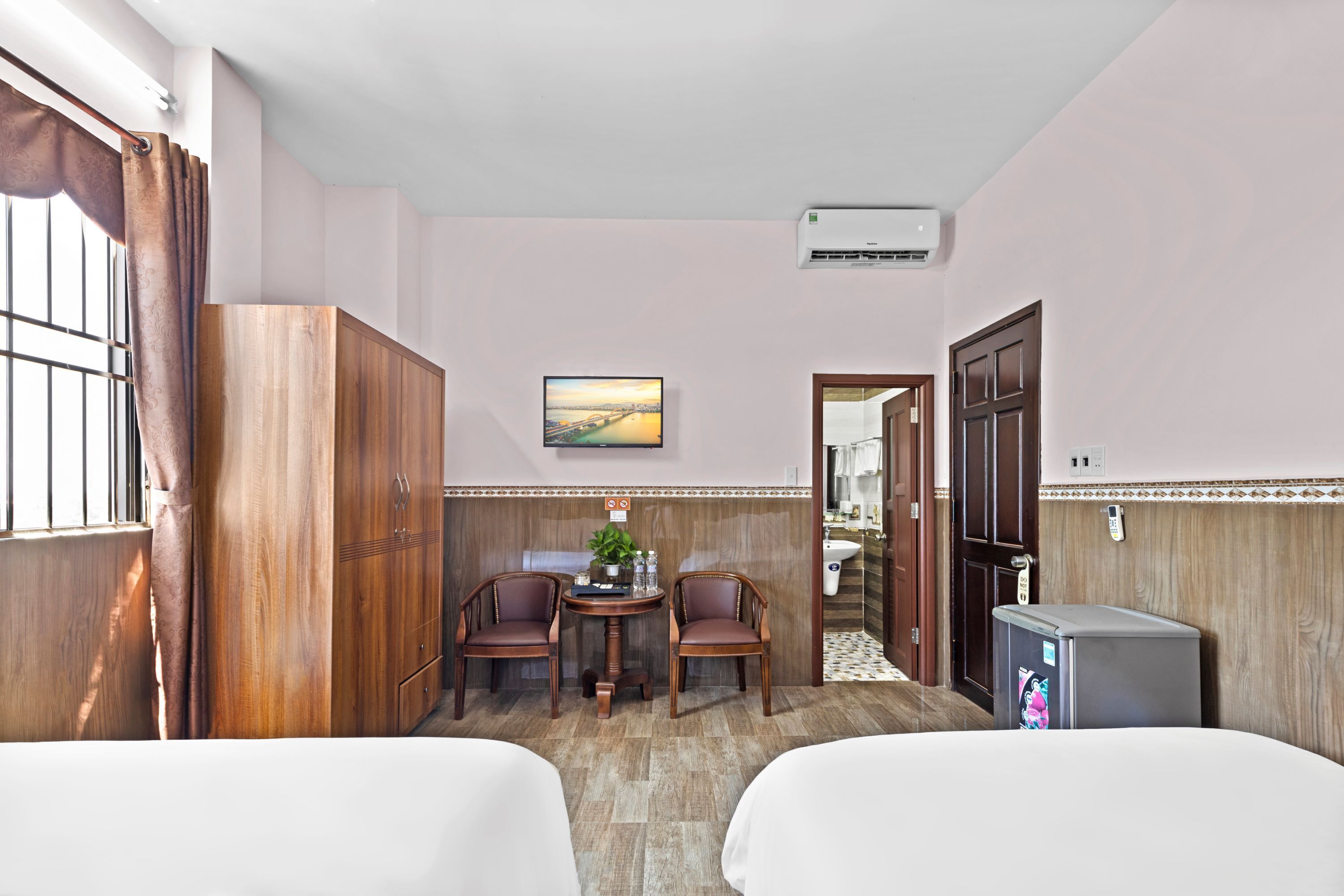 A hotel room with two beds, a wooden wardrobe, a small table with chairs, a painting on the wall, and an air conditioning unit.