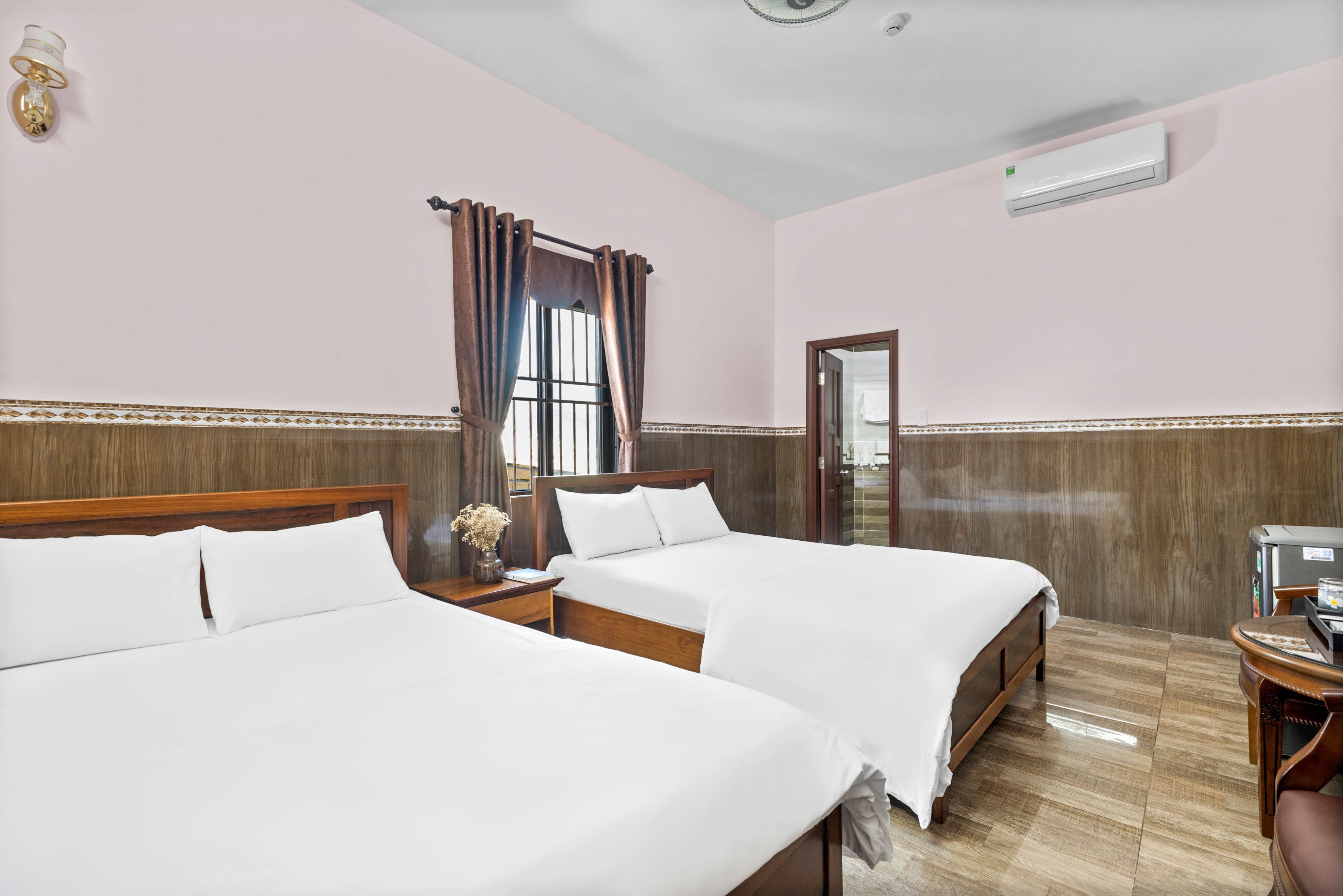 A hotel room with two beds, a wooden headboard, a small table with a lamp, and an air conditioning unit on the wall.