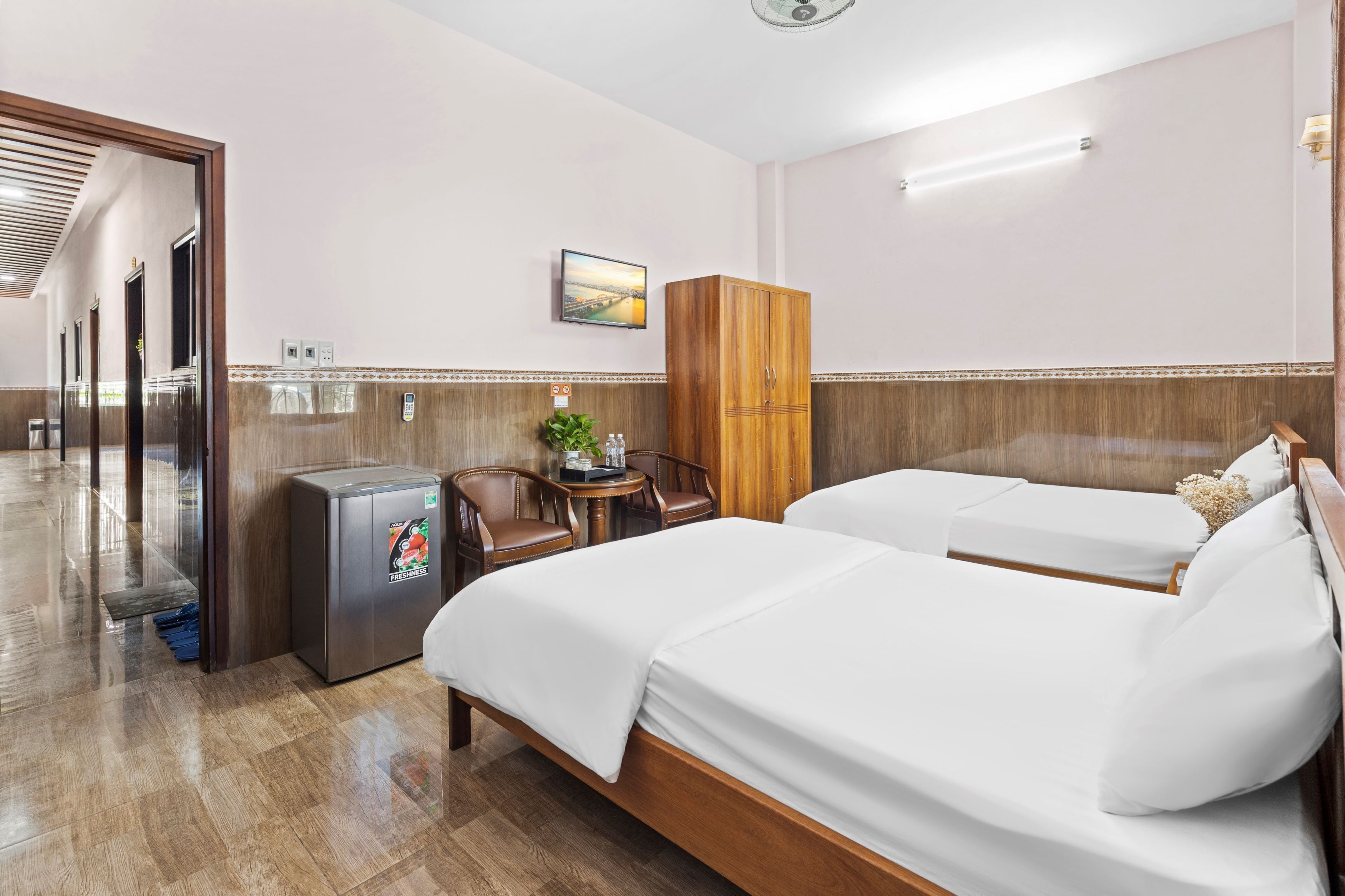 A hotel room with two beds, a wooden wardrobe, a small table with chairs, a refrigerator, and a wall-mounted TV.