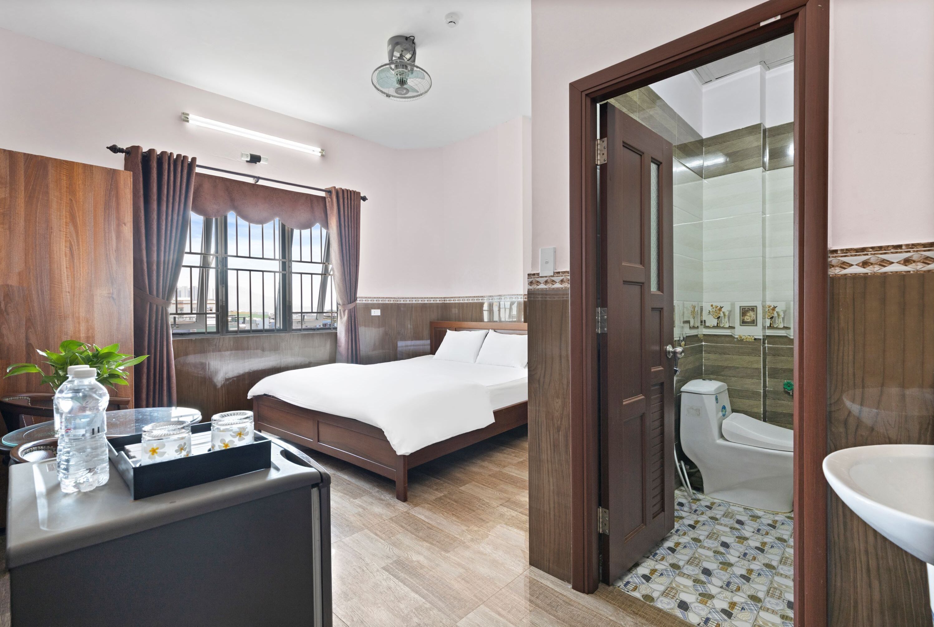 A hotel room with a bed, bedside table with a bottle and glasses, a window with curtains, a bathroom with a toilet and sink, and a door leading to another room.