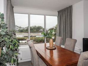 A dining area with a wooden table, grey upholstered chairs, a vase with white flowers, and a view of a coastal landscape through large windows. - Ivy Cottage (Porthmadog)