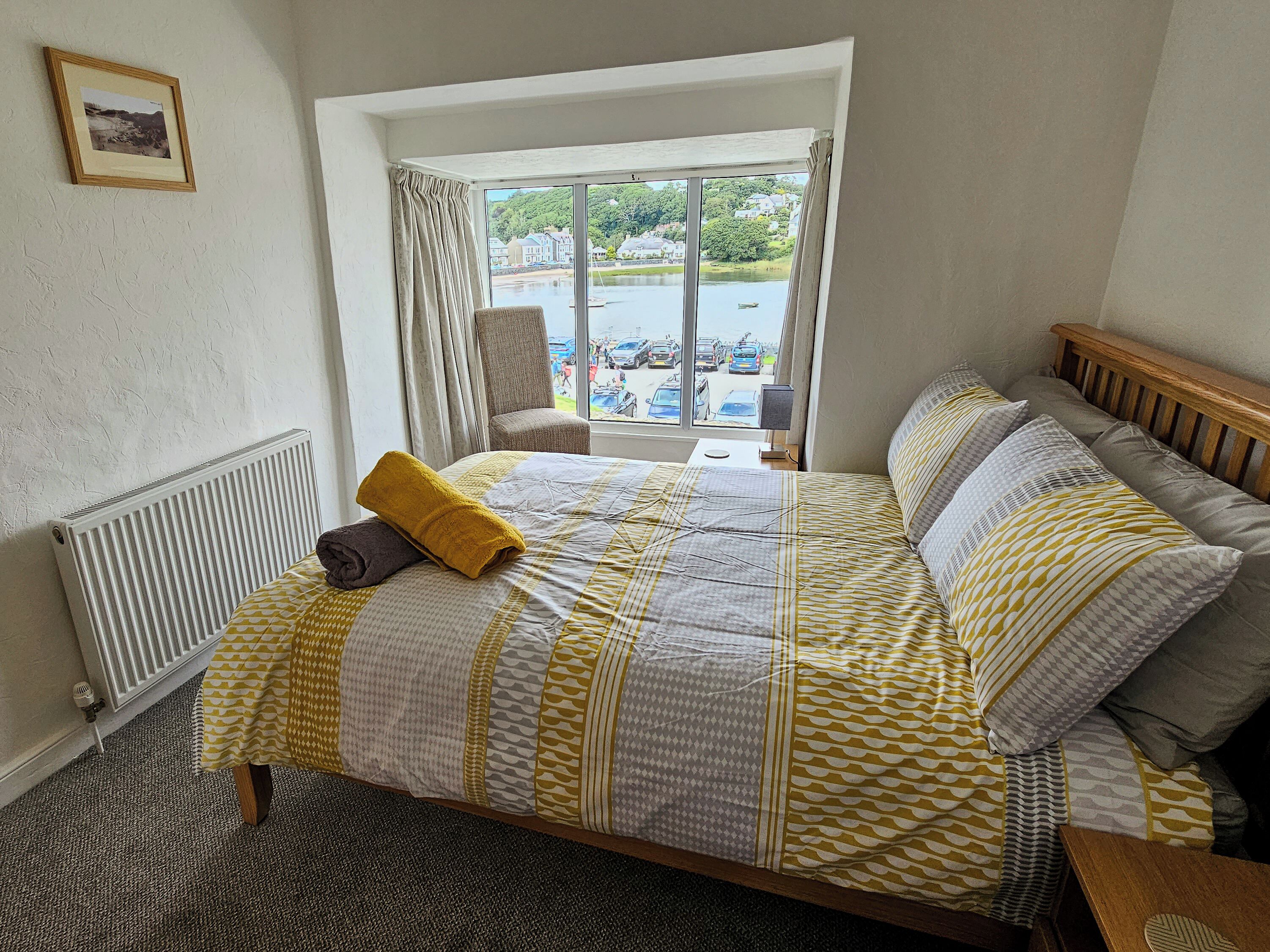 A bedroom with a large window, a bed with patterned bedding, a chair, and a radiator.