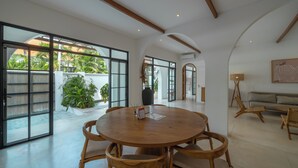 A spacious room with a round wooden table and chairs, large windows, and a view of greenery outside. - Villa Eliad - 3 Bedroom Villa in Berawa (Canggu)