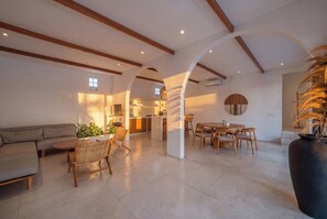 A spacious living area with a sofa, dining table, and chairs, a kitchen in the background, and a large plant.