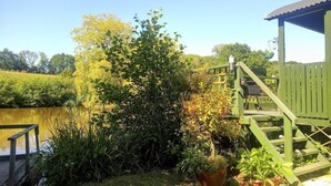 A wooden deck with a metal railing, a green gazebo, and a pond with surrounding vegetation. - Cosy, peaceful & secluded lakeside cabin. BBQ, Firepit, Fishing, Dogs welcome. (Hadlow Down)