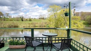 A deck with two chairs and a table overlooking a body of water, with a lamp post and hanging lamps. - Cosy, peaceful & secluded lakeside cabin. BBQ, Firepit, Fishing, Dogs welcome. (Hadlow Down)