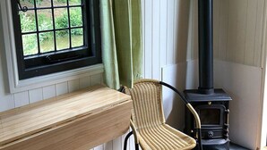 A room with a wooden bench, a wicker chair, a window with a view of greenery, and a black wood-burning stove. - Snug secluded lakeside shepherds hut for fishing, or just relaxing.  (Hadlow Down)