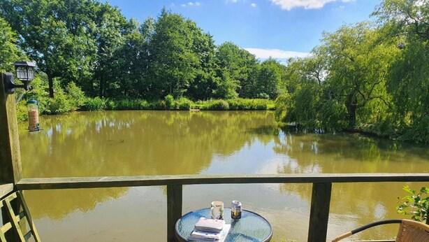 A wooden deck with two wicker chairs and a small round table with a book and a can. - Snug secluded lakeside shepherds hut for fishing, or just relaxing.  (Hadlow Down)