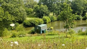 A serene lakeside setting with a small shed, lush greenery, and a calm water body. - Snug secluded lakeside shepherds hut for fishing, or just relaxing.  (Hadlow Down)