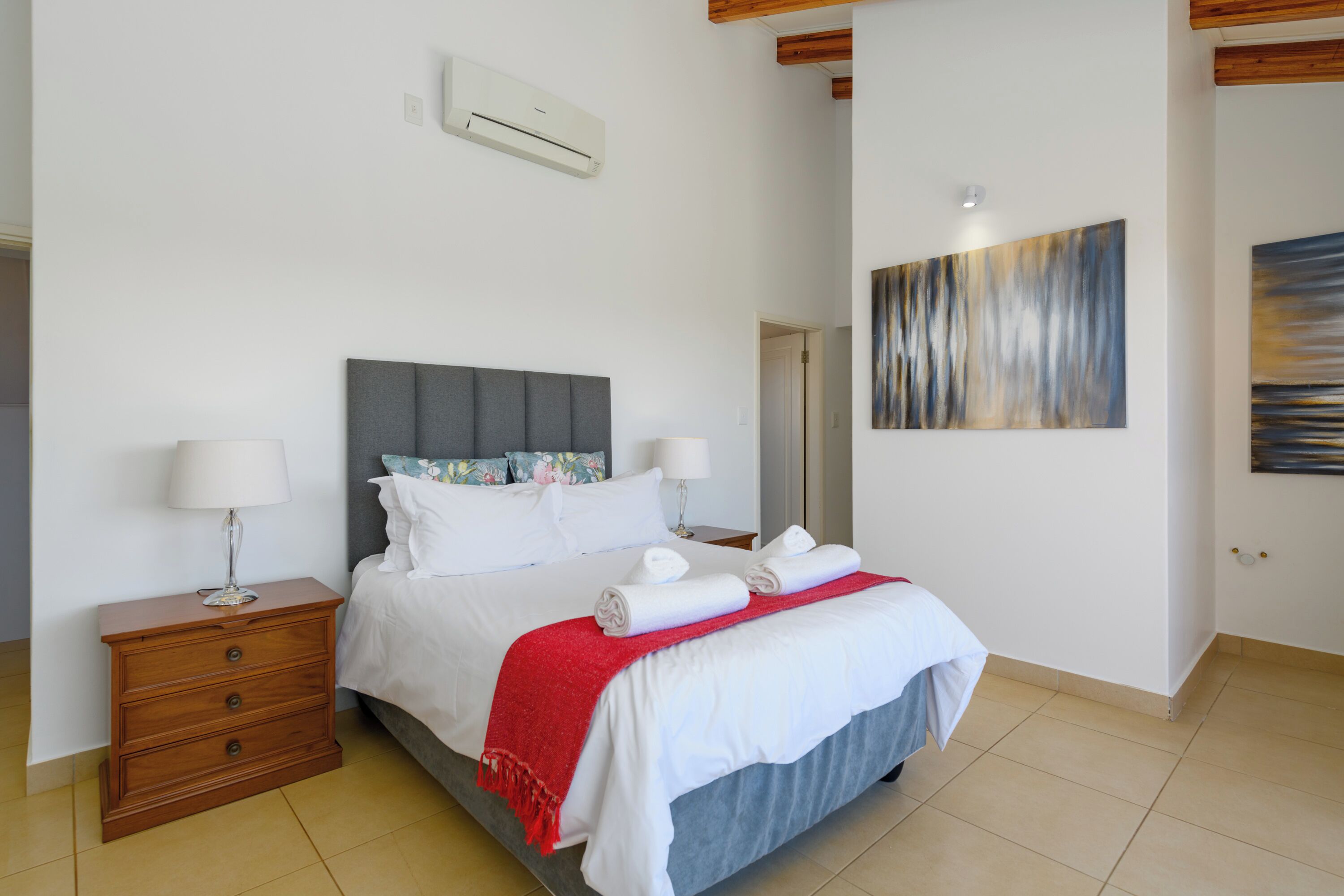 A neatly made bed with white linens and a red throw blanket, a wooden nightstand with a lamp, and a modern abstract painting on the wall.