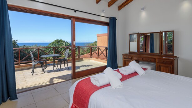 A bedroom with a large bed, a wooden dresser, a mirror, and a balcony with a table and chairs. - San Lameer Villa 14323 (Southbroom)