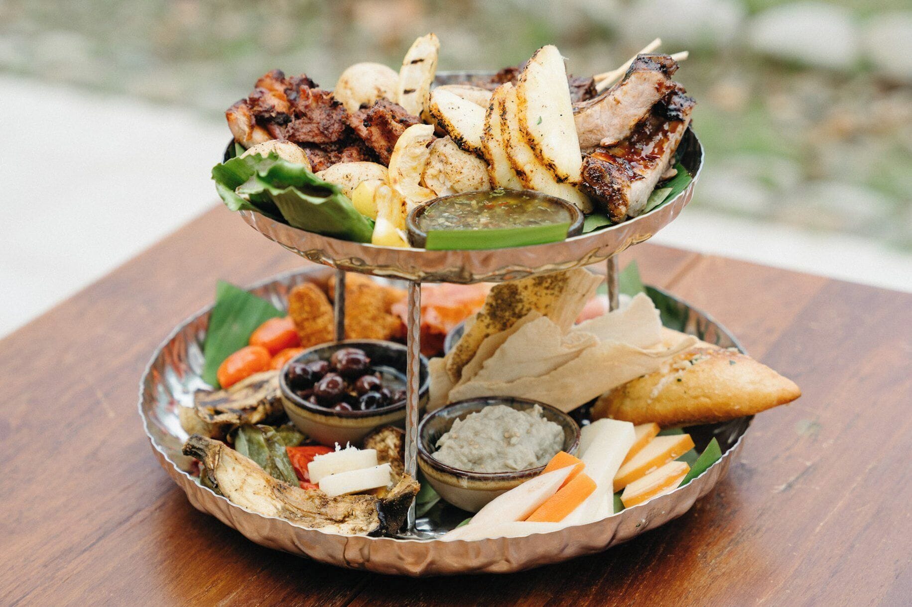 A multi-tiered serving platter with various foods including grilled meats, potatoes, and dips.