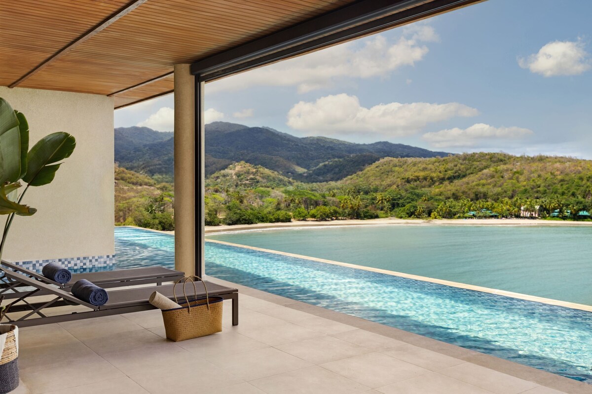 A poolside area with lounge chairs and a view of a beach and mountains.