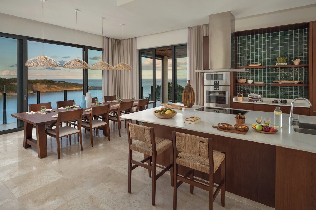 A modern kitchen with a large island, dining area with chairs, and a view of the sea.
