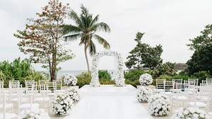Un pasillo nupcial con sillas blancas y arreglos florales, que conduce a un arco.