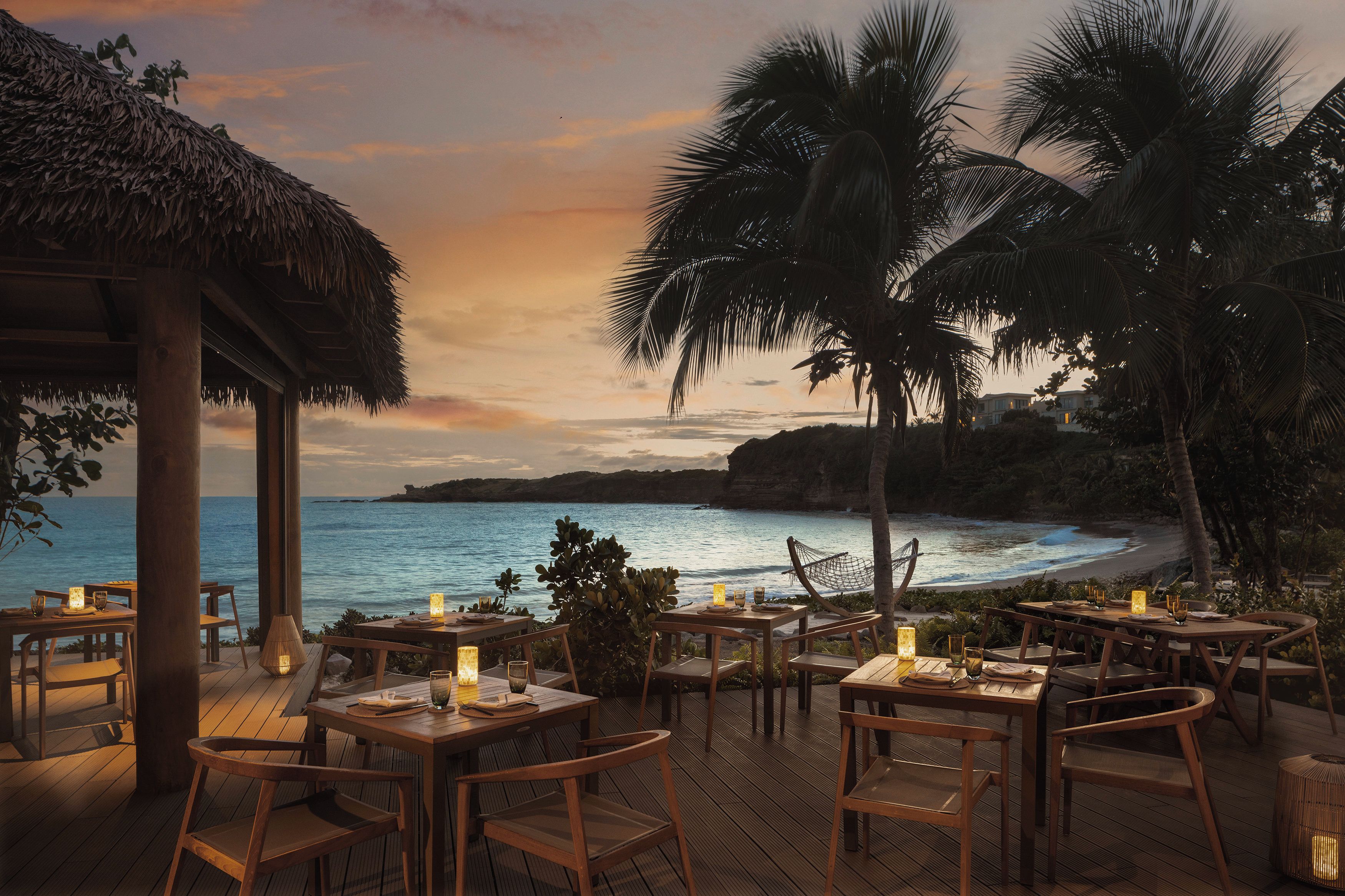 A seaside restaurant with wooden tables and chairs, a thatched roof, and a view of the ocean.