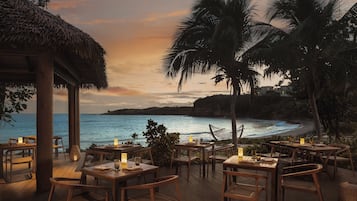 A seaside restaurant with wooden tables and chairs, a thatched roof, and a view of the ocean.