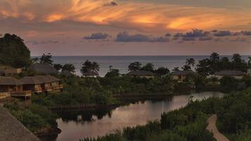 A resort with thatched-roof bungalows, a calm lake, and a view of the ocean at sunset.