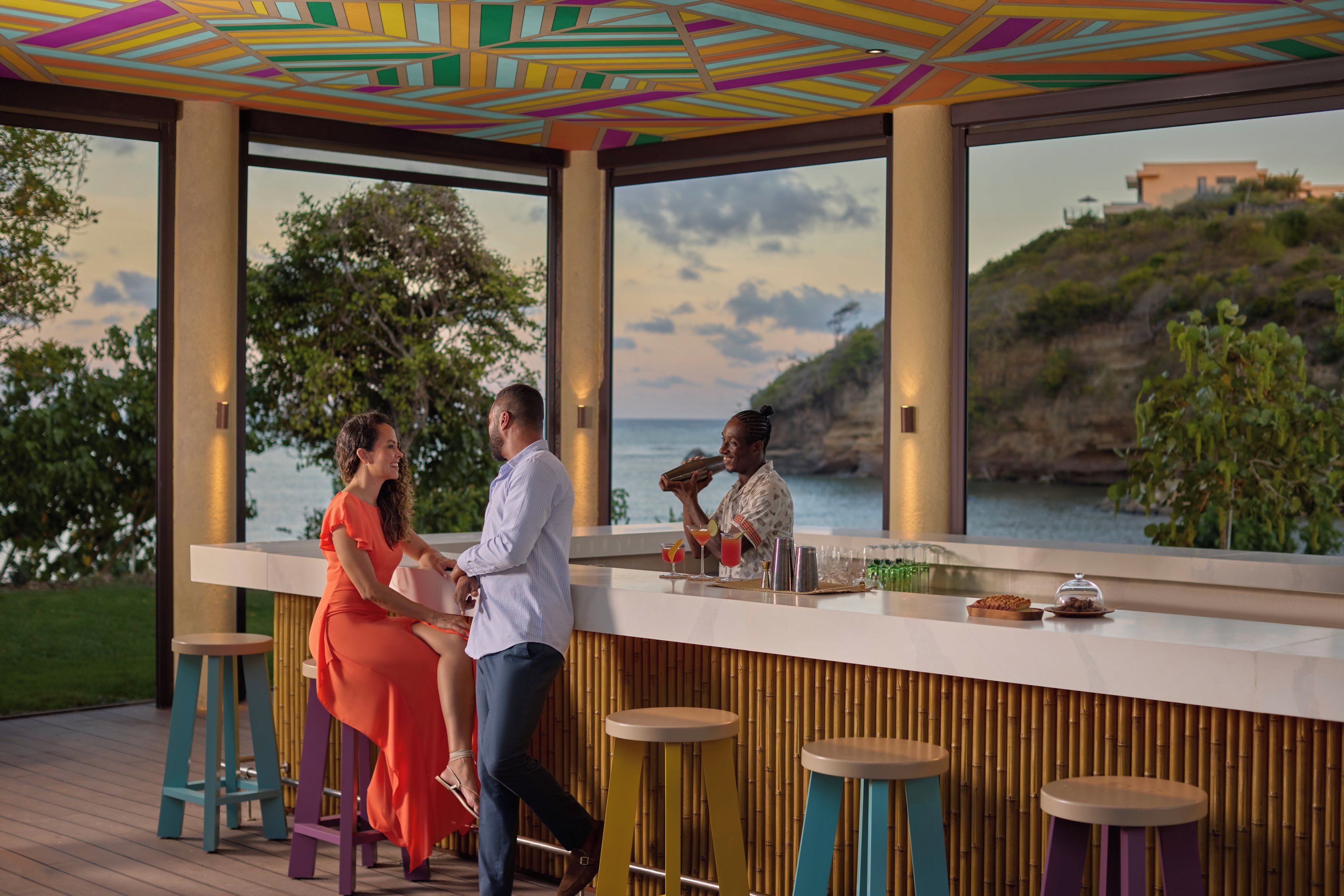 Zona de bar con vista al mar, paredes de bambú y taburetes coloridos.