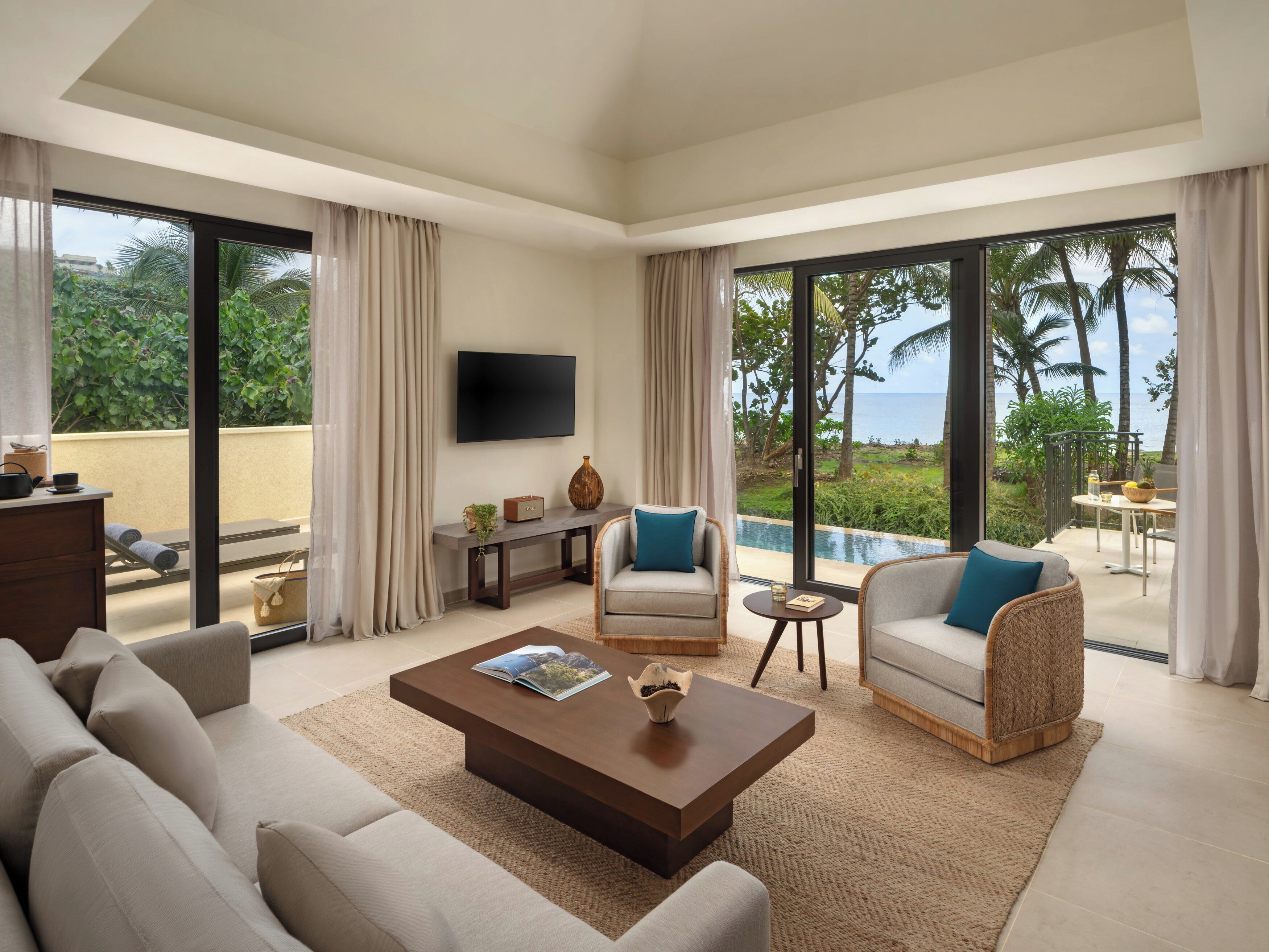 A modern living room with a sofa, armchairs, a coffee table, and a TV. There is a view of the ocean through large windows.