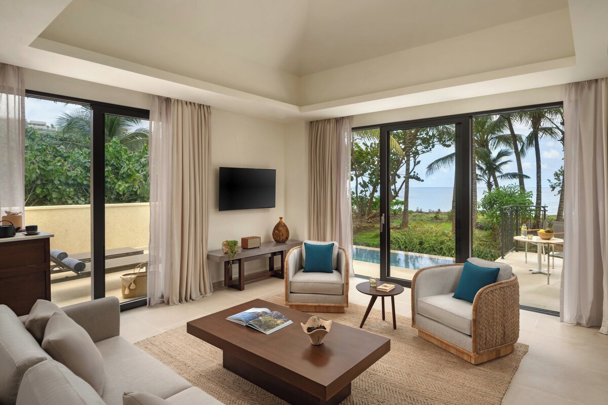 A spacious living room with a large sofa, a coffee table, and a TV mounted on the wall. There is a view of a pool and palm trees through large windows.
