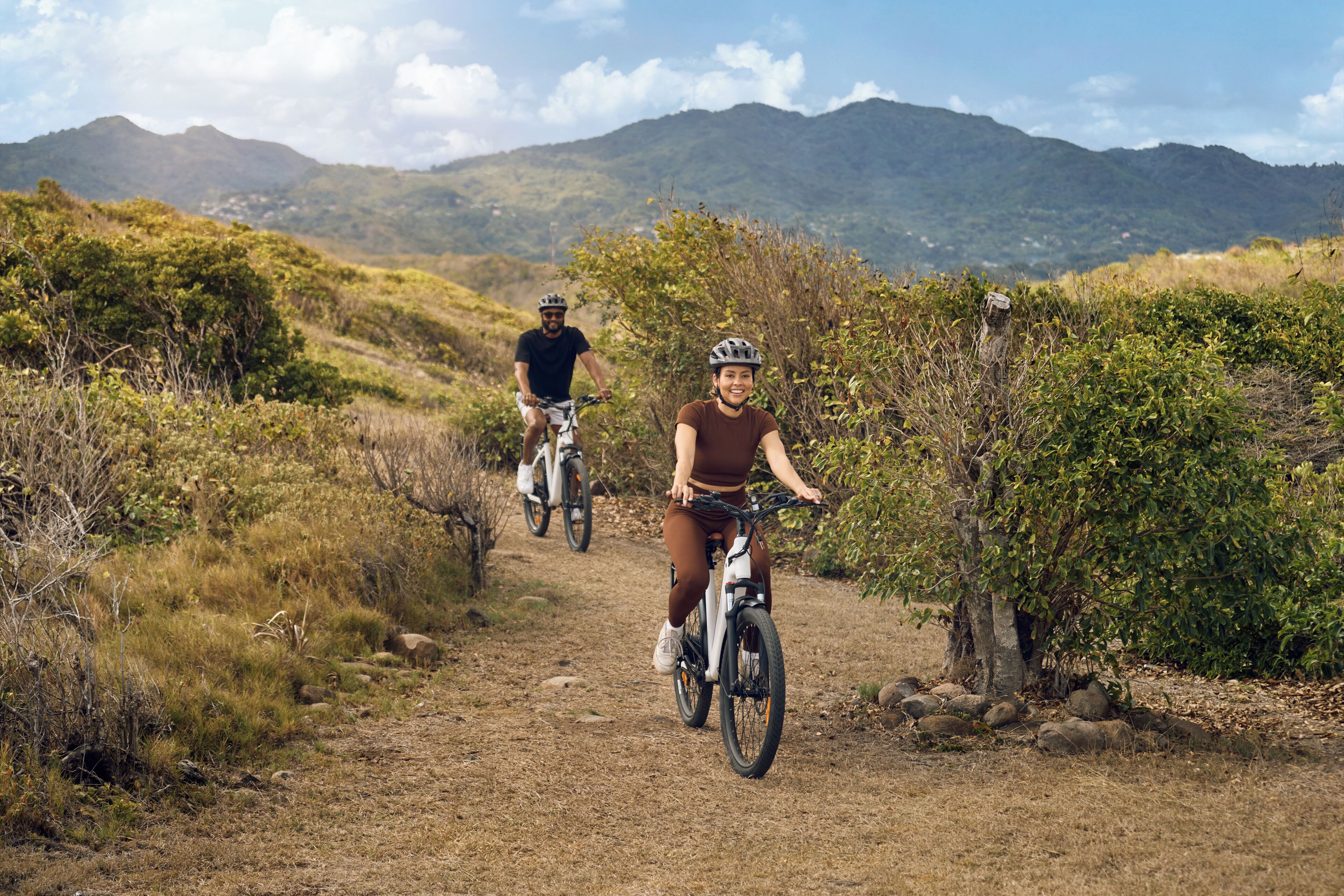 Two people riding bicycles on a dirt path surrounded by greenery and mountains.