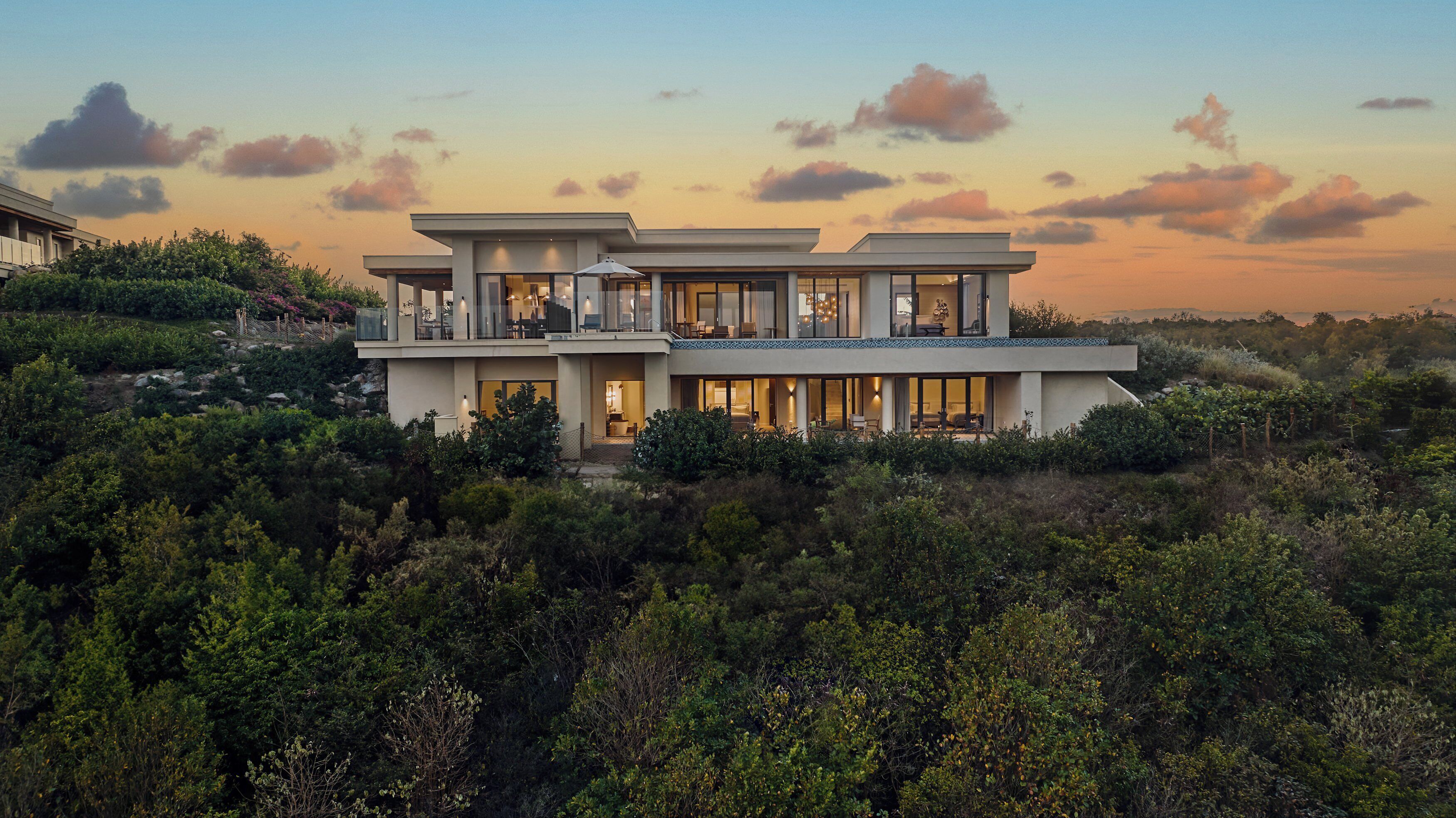 Et moderne hus i to etager med en balkon, omgivet af frodig vegetation.
