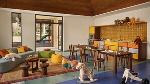 Amplia sala de estar con sofá, mesa de centro y zona de juegos. Se observa una cocina con mesa y sillas.